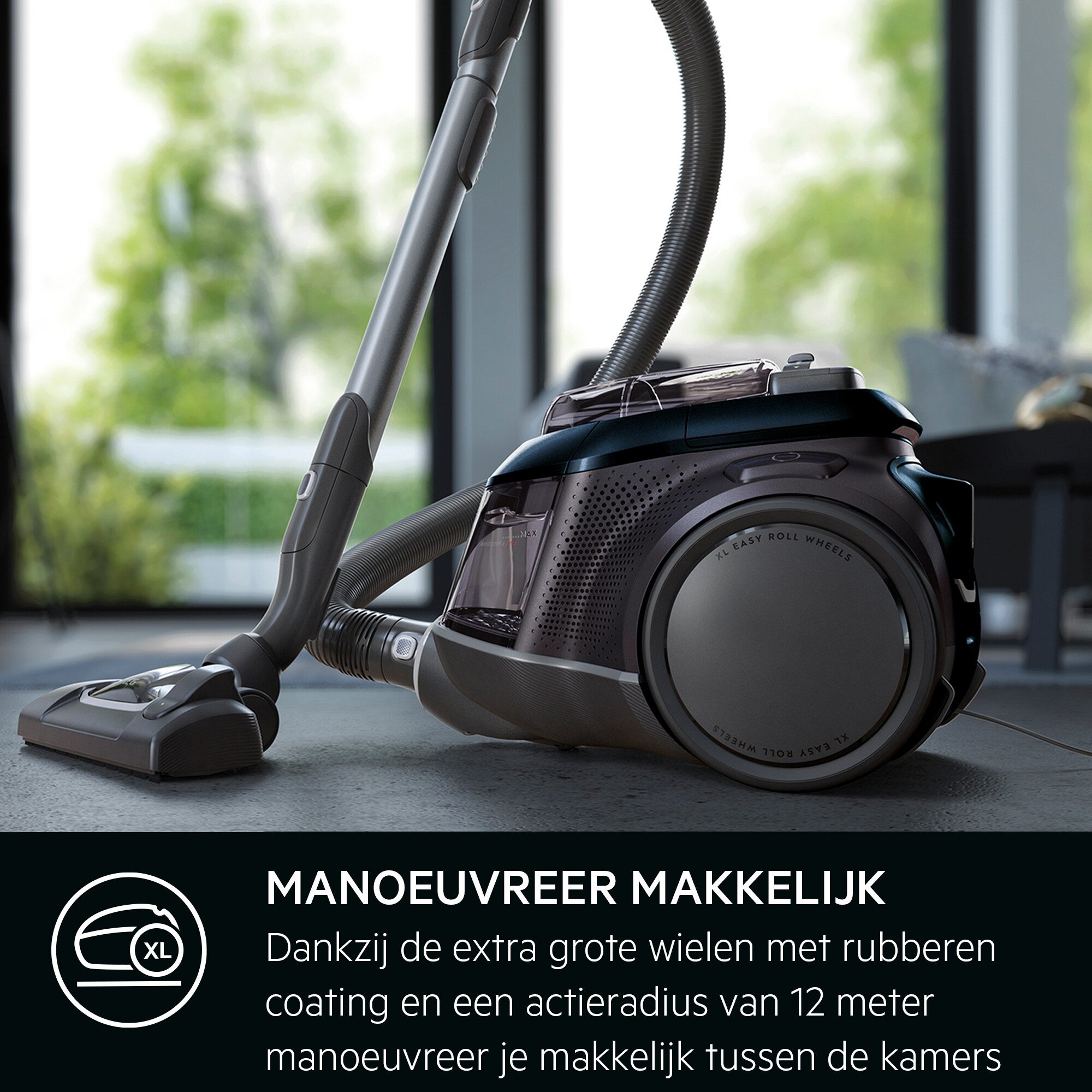 Un aspirateur sombre sur un sol gris. Il a une longue poignée et une grande roue avec 'XL EASY ROLL WHEELS'.