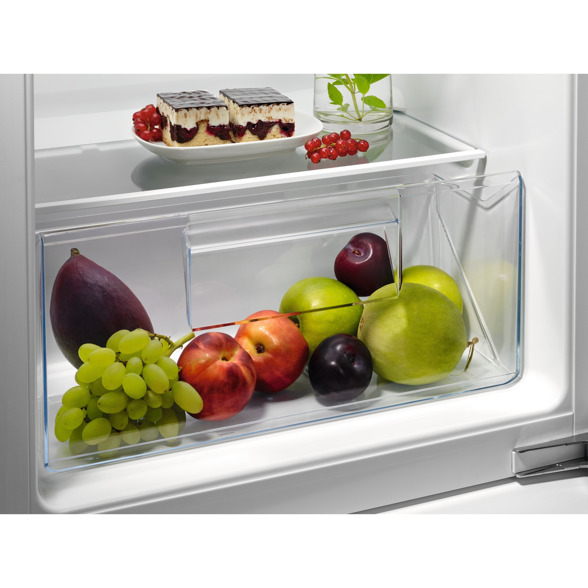 In einem Kühlschrank ein Teller mit Kuchen und roten Beeren und eine Schublade voller Früchte: Trauben, Pflaumen, Äpfel und Birnen.
