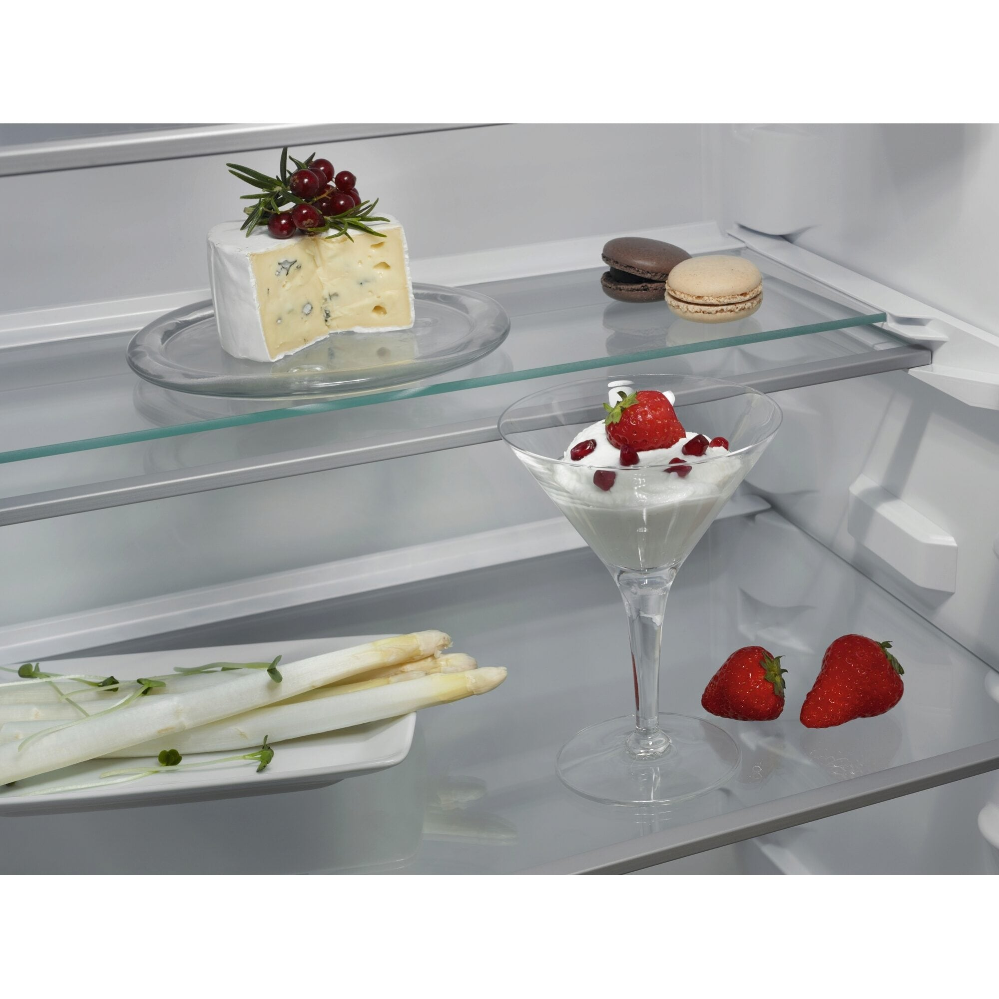 Innenraum eines Kühlschranks mit Käse, Makronen, Spargel und einem Dessert im Glas. Erdbeeren sind ebenfalls vorhanden.