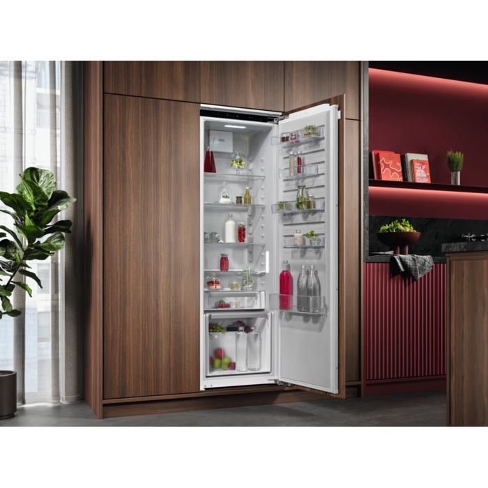 Ein offener Kühlschrank in einem Holzschrank, gefüllt mit Lebensmitteln. Im Hintergrund sind eine Zimmerpflanze und eine rote Wand zu sehen.