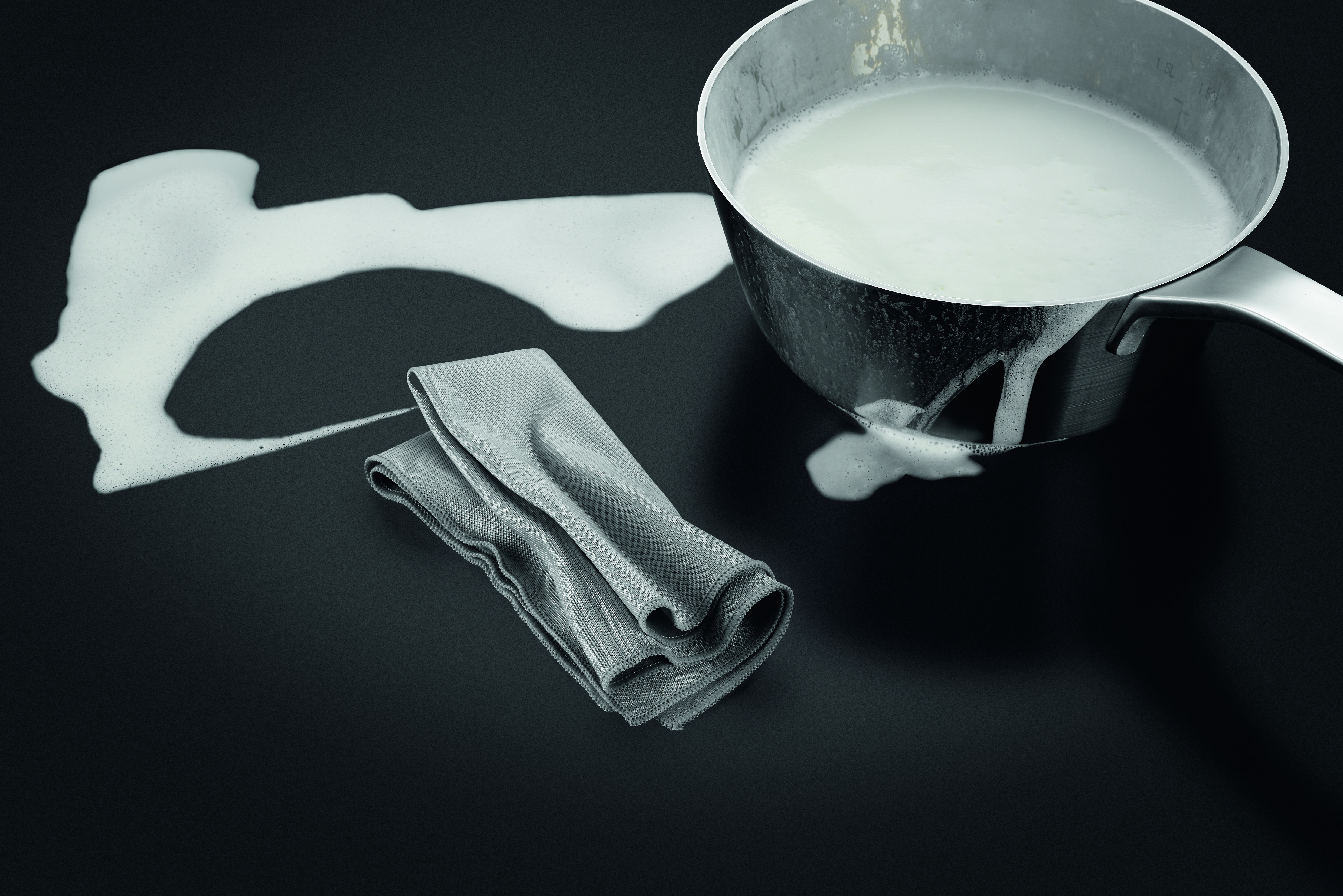 Ein Topf mit Milch, die auf eine schwarze Oberfläche verschüttet wird, neben einem gefalteten grauen Tuch. Schaum breitet sich aus.