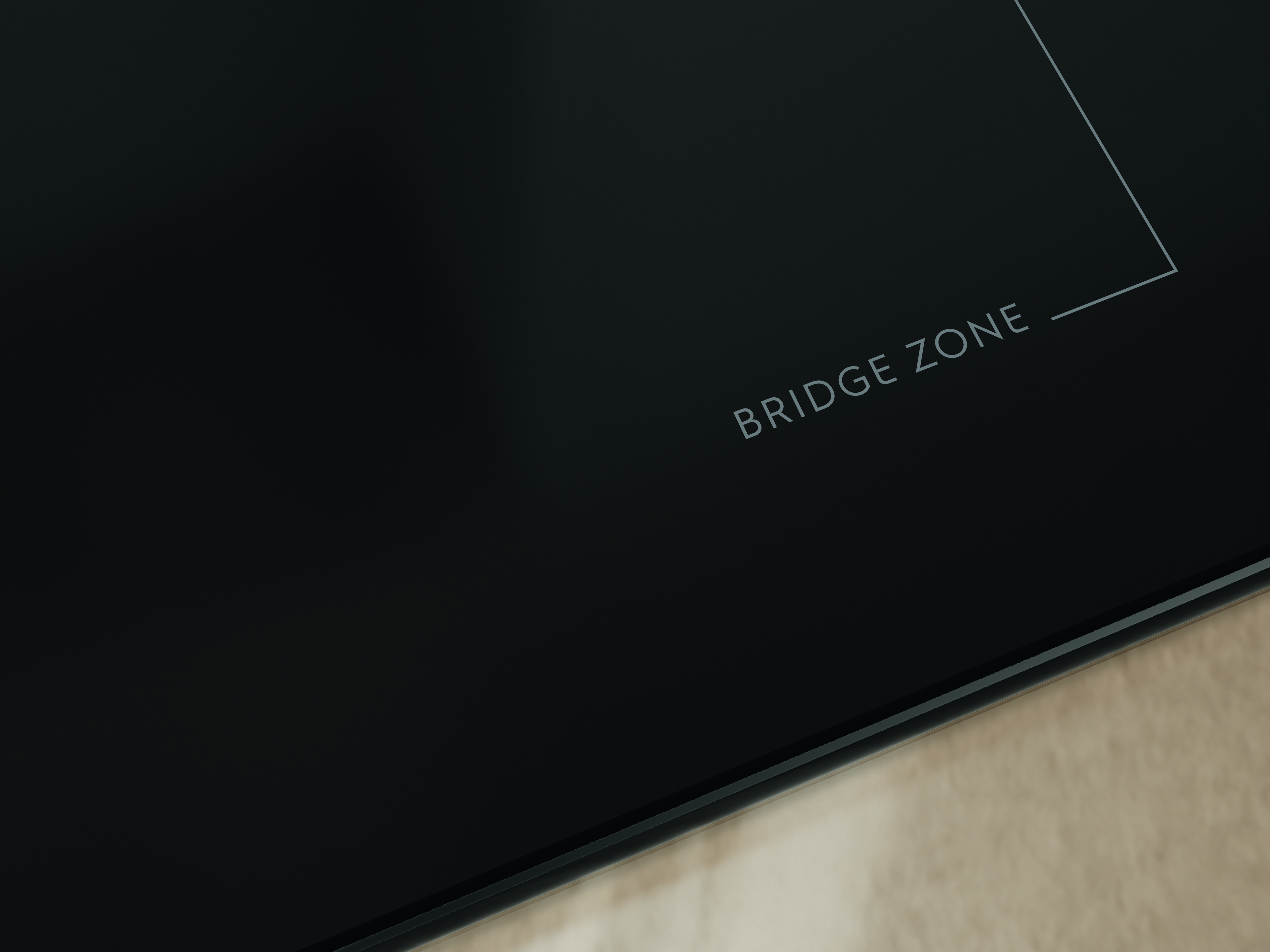 Nahaufnahme eines schwarzen Kochfelds mit dem Text 'BRIDGE ZONE' und einer weißen Linie, die einen Bereich markiert. Helle holzähnliche Oberfläche unten.