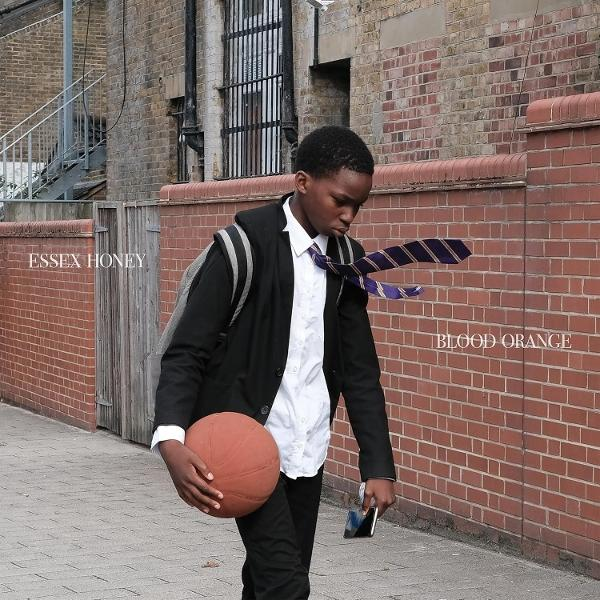 Ein junger Mann in Uniform geht mit einem Basketball und Telefon. Im Hintergrund sind eine Backsteinmauer und ein Gebäude.