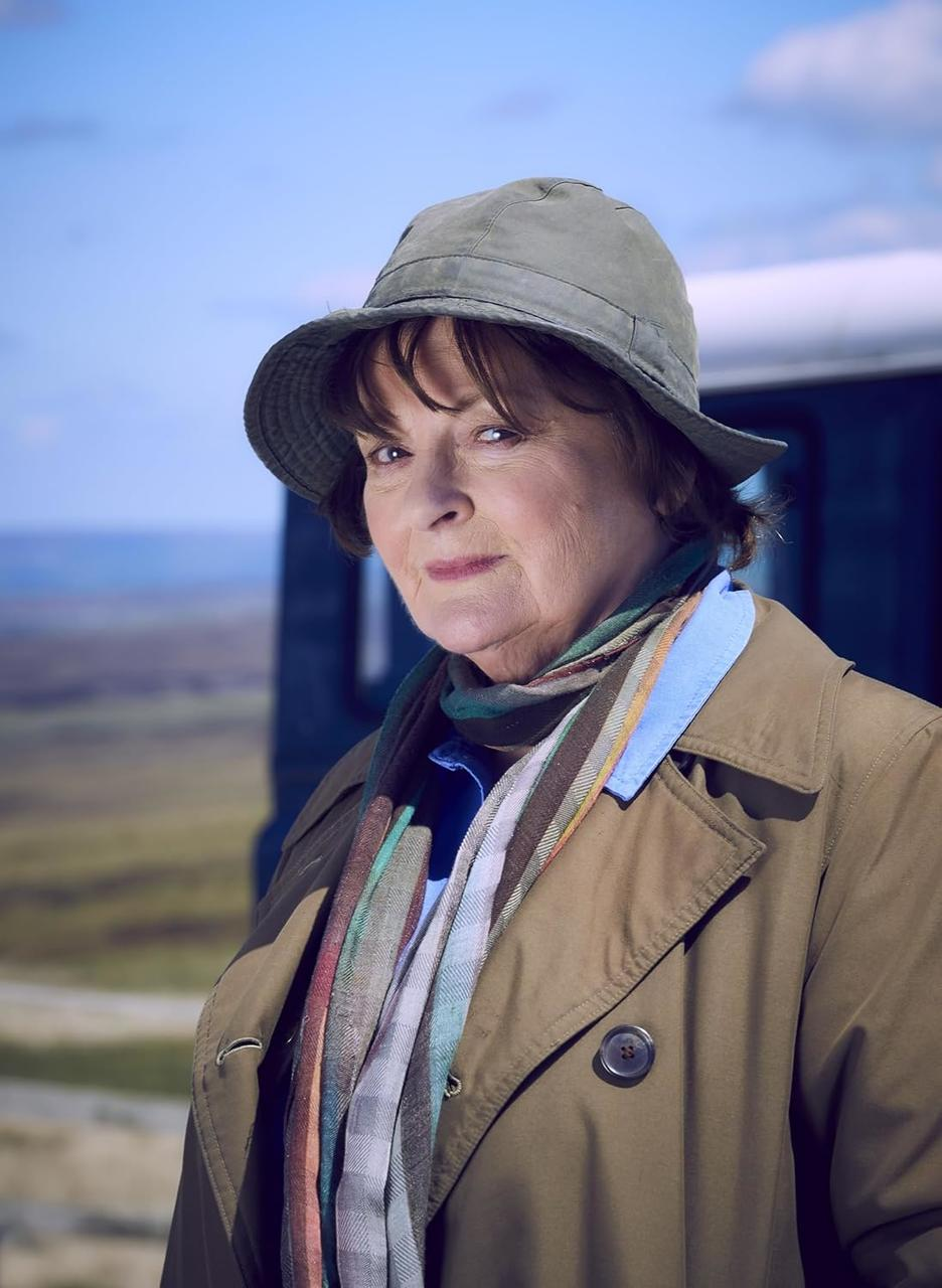 Eine Frau mit Hut, Mantel und Schal steht im Freien. Der Hintergrund zeigt eine Landschaft mit blauem Himmel und einem Fahrzeug.