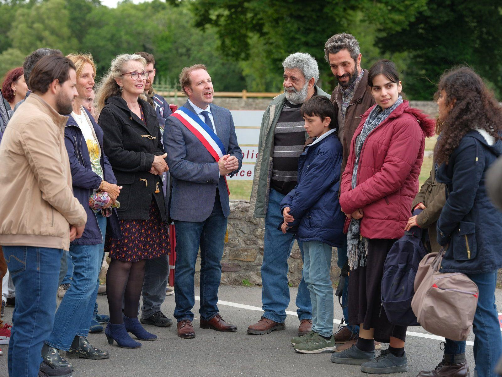 Eine Gruppe von Menschen steht und unterhält sich im Freien. Ein Mann trägt eine Schärpe. Bäume im Hintergrund.