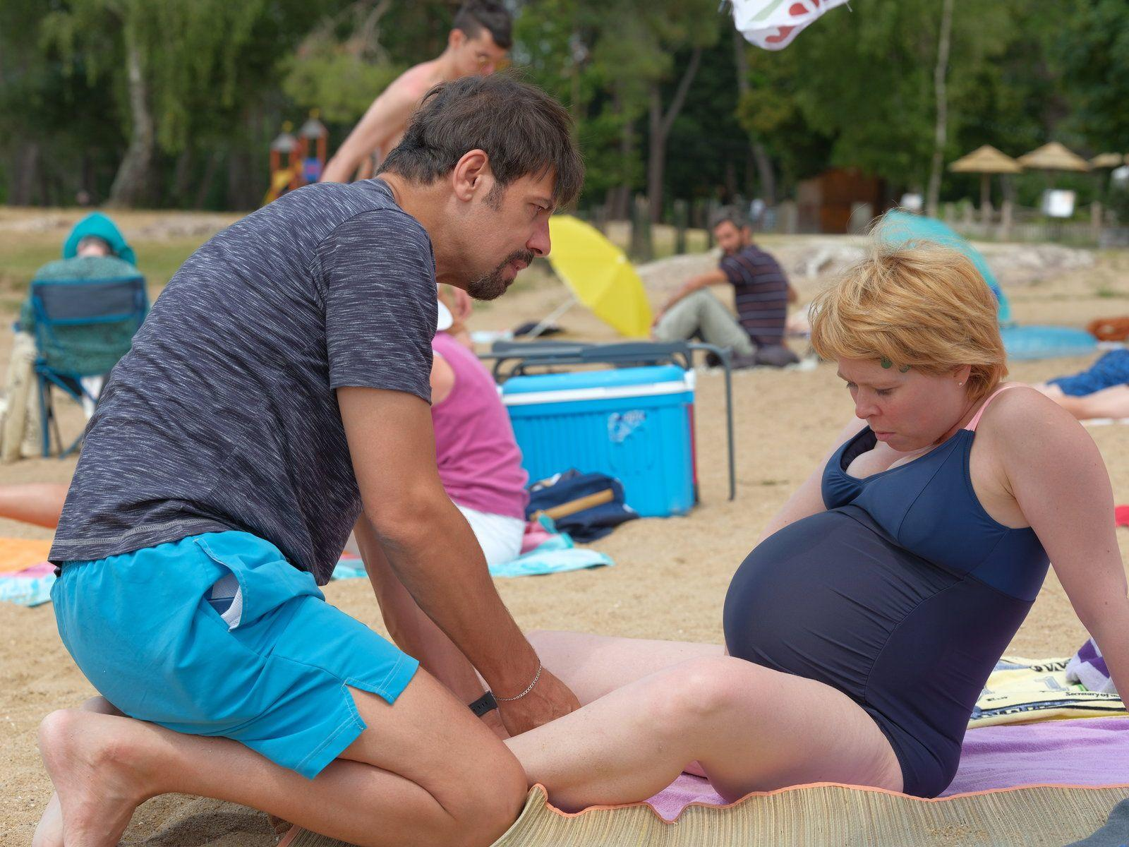 Ein Mann hilft einer schwangeren Frau am Strand. Sie sitzt auf einem Handtuch. Andere Leute sind im Hintergrund.