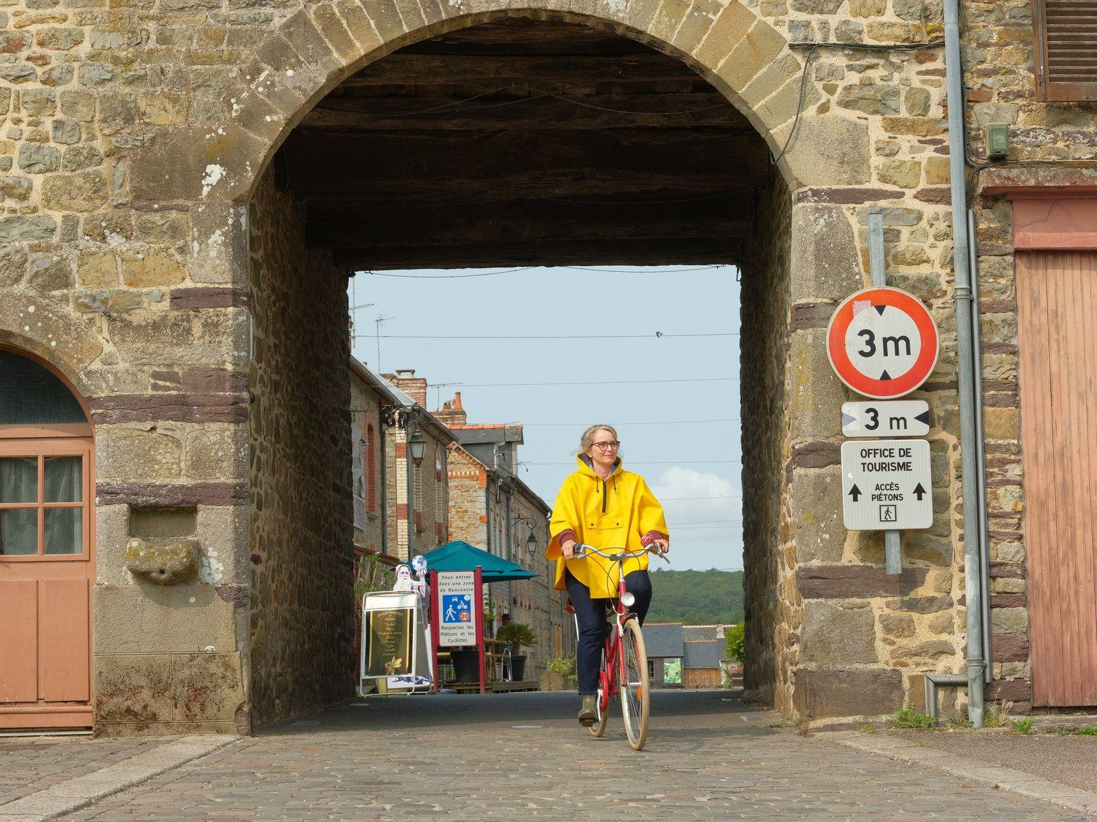 Frau im gelben Regenmantel fährt mit dem Fahrrad durch einen Steinbogen. Schilder weisen auf 3 m Durchfahrtshöhe hin.