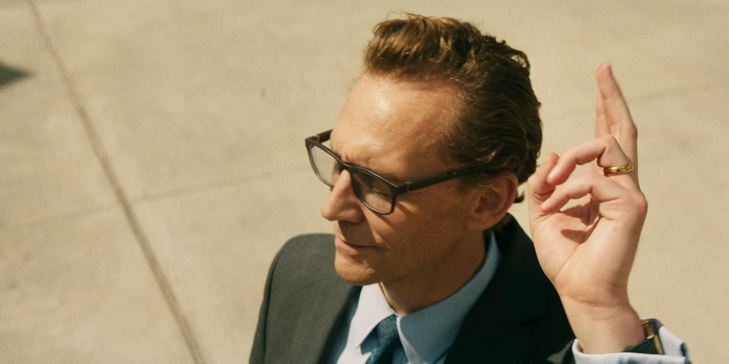 Mann mit Brille, Anzug und Ring, der ein Okay-Zeichen macht. Beiger Hintergrund. Fokus auf Gesicht und Handgeste des Mannes.