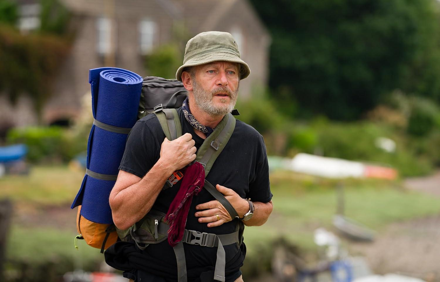 Ein Mann mit Hut und Rucksack geht im Freien. Er trägt eine blaue Matte. Im Hintergrund sind ein Gebäude und grüne Vegetation zu sehen.
