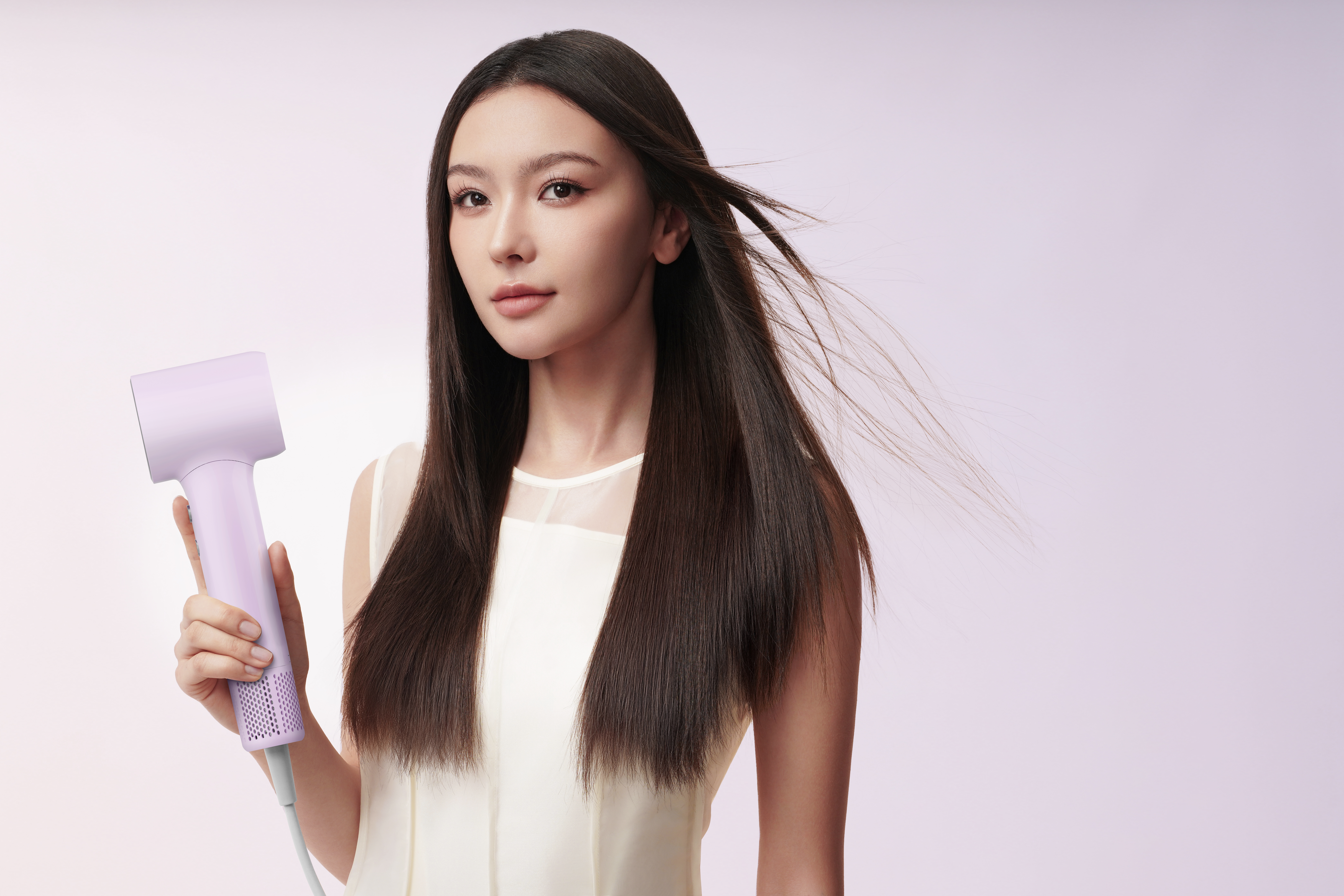 Kobieta z długimi, ciemnymi włosami trzyma liliową suszarkę do włosów. Ma na sobie biały top. Tło jest jasno-różowe.