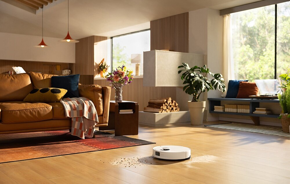 Salon z brązową skórzaną sofą, robotem sprzątającym, oknem i roślinami. Podłoga jest drewniana.