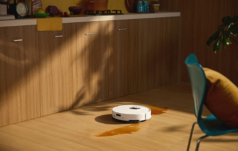 Biały robot sprzątający na drewnianej podłodze. Brązowe plamy wokół niego. Niebieskie krzesło z żółtą poduszką po prawej.