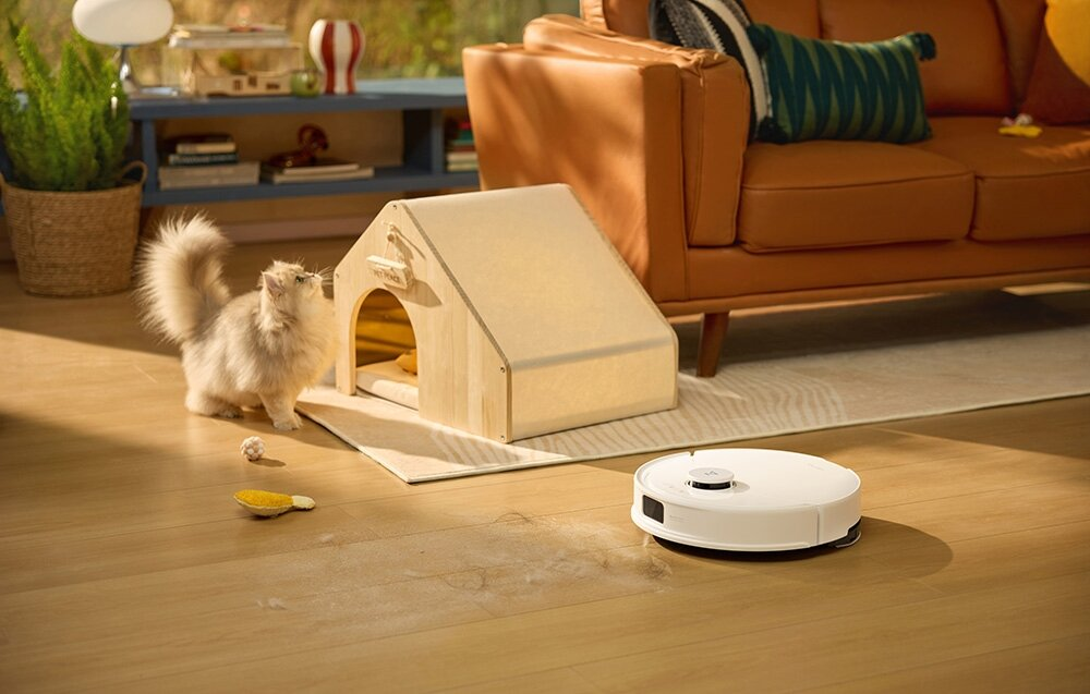 Puszysty kot patrzy na drewniany domek dla kota. Biały odkurzacz robot sprząta drewnianą podłogę. Brązowa sofa jest w tle.