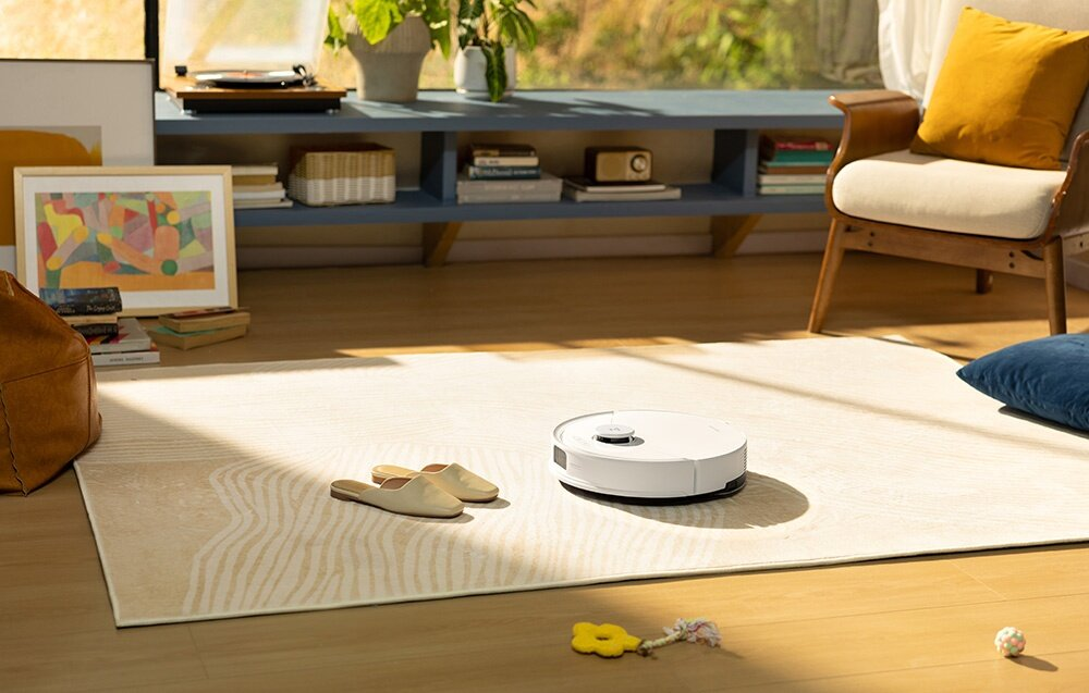 Biały odkurzacz robot na beżowym dywanie. Kapcie, żółty klucz i krzesło na pierwszym planie. Okno w tle.