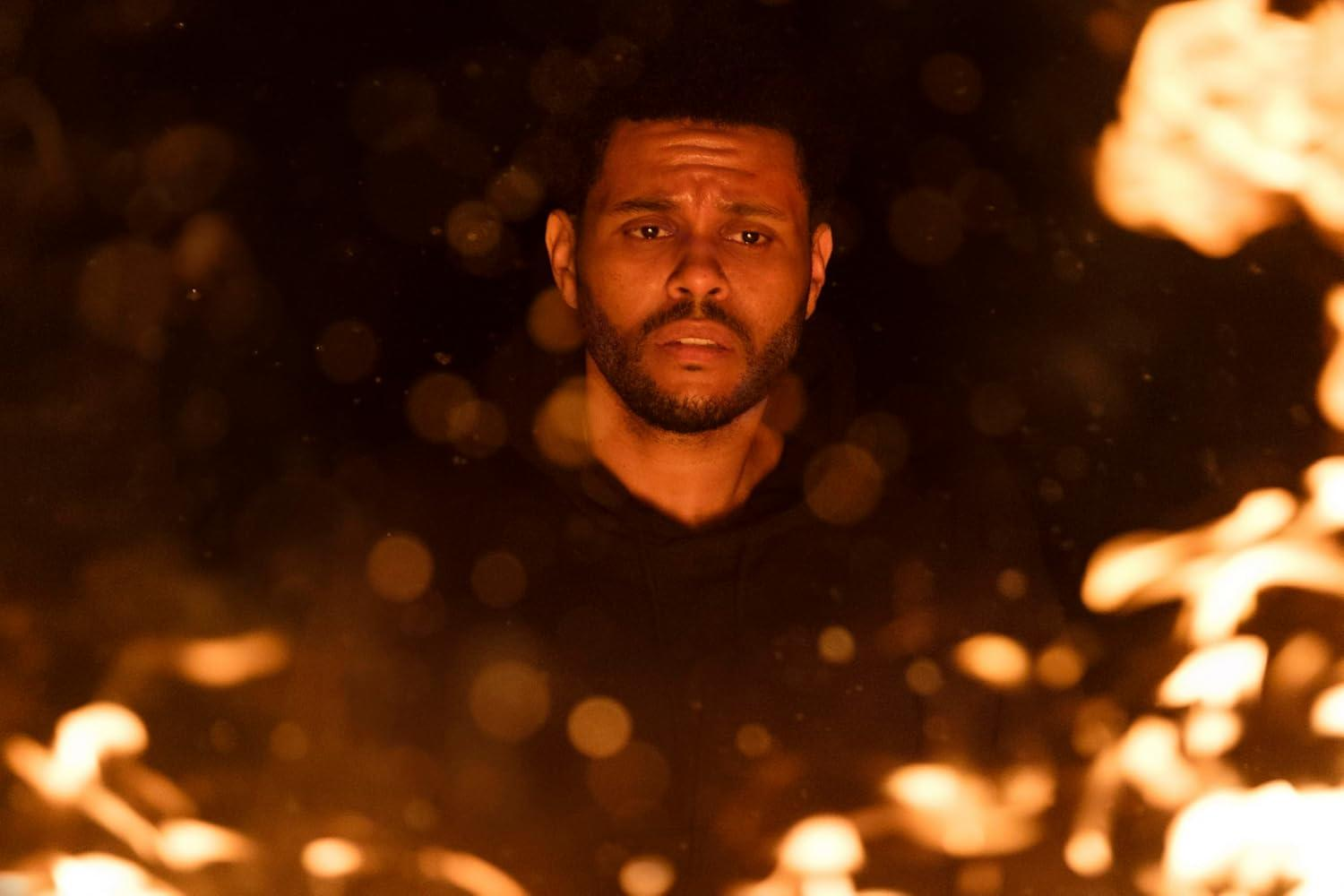 Ein Mann mit Bart blickt in die Kamera. Flammen umgeben ihn und erzeugen ein orangefarbenes Leuchten. Dunkler Hintergrund mit Bokeh.