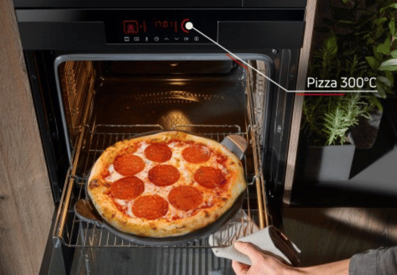 Pizza pepperoni piecze się w piekarniku ustawionym na 300°C. Drzwi piekarnika są otwarte, a ręka używa łopatki do pizzy.