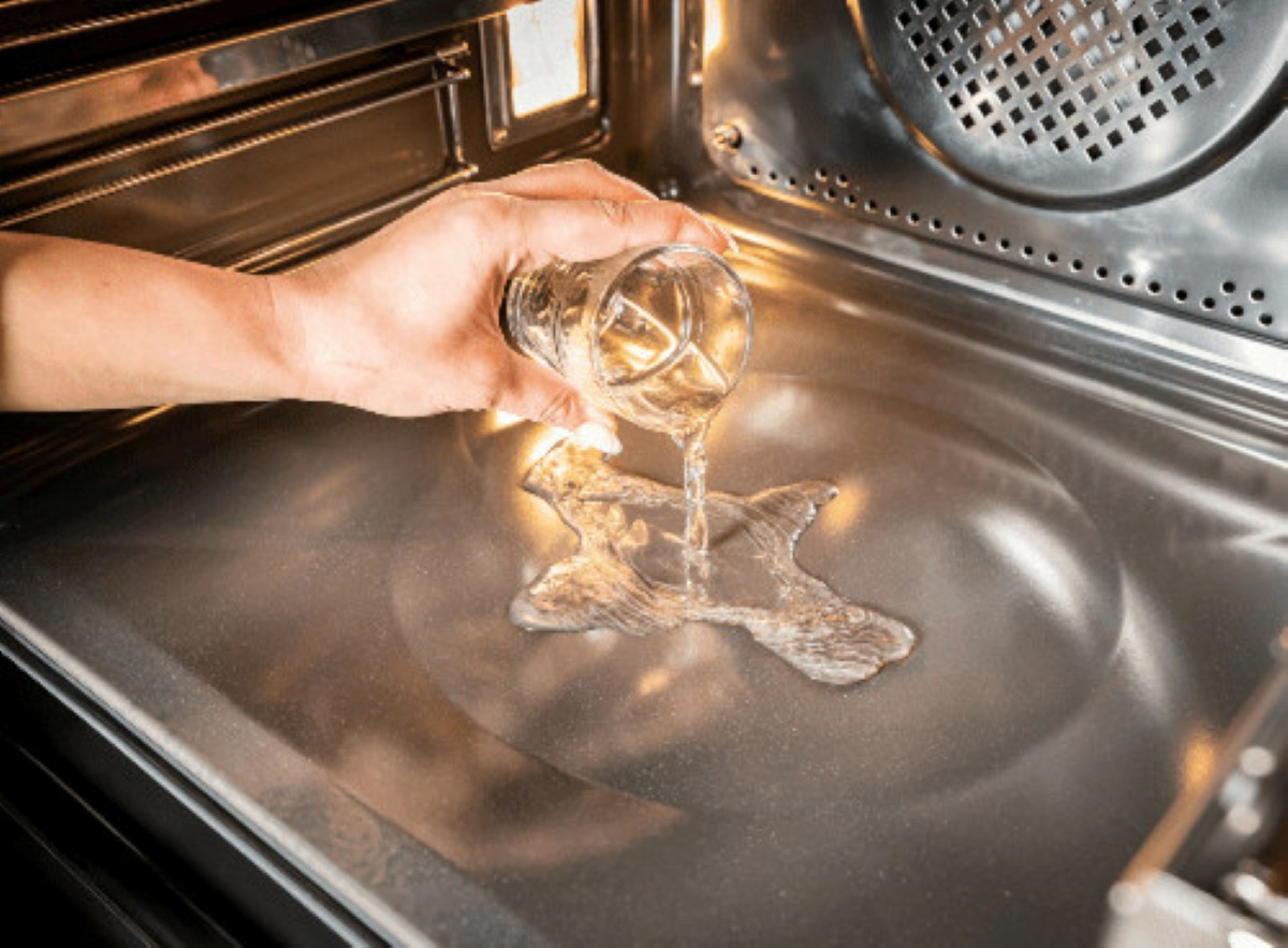 Osoba wlewa wodę ze szklanki do piekarnika. Woda tworzy kałużę na dnie piekarnika. Piekarnik jest ze stali nierdzewnej.