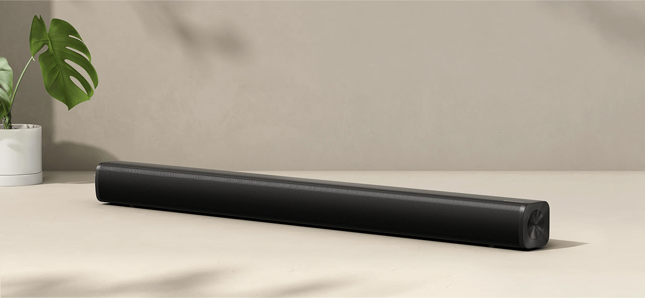 Czarny soundbar na jasnej powierzchni. Zielona roślina po lewej. Tło to jasnobeżowa ściana.