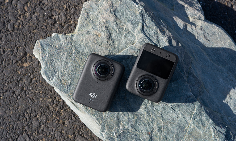 Dwie czarne kamery sportowe leżą na szarym kamieniu. Jedna ma ekran, druga obiektyw. Tło to ciemny asfalt.