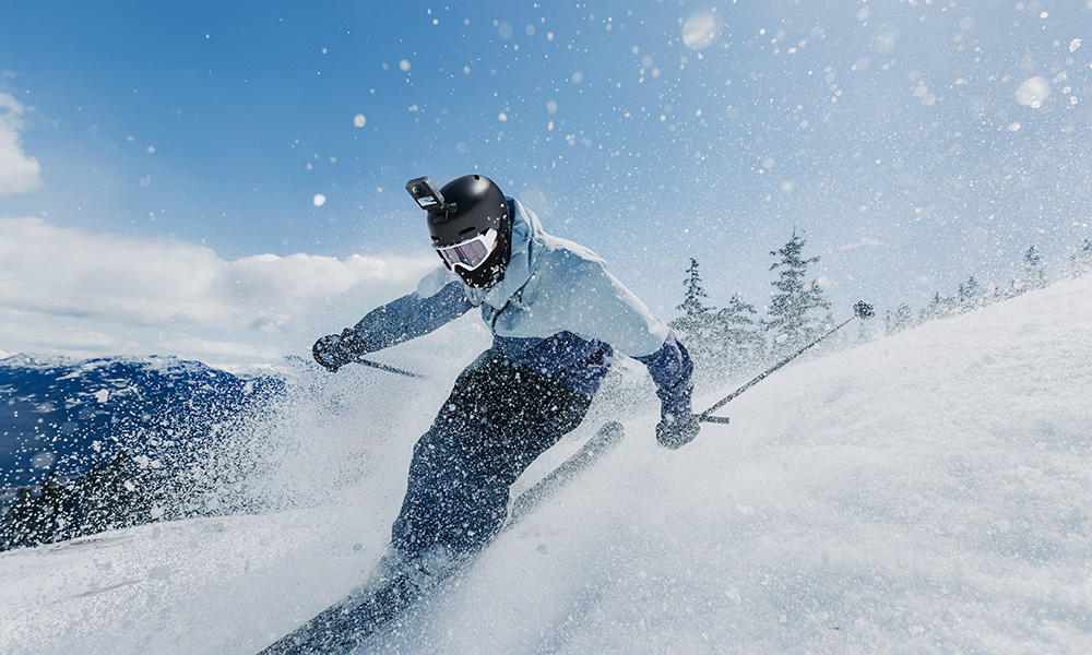 Narciarz zjeżdża ze śnieżnego stoku. Ma kask z kamerą, gogle i jasno niebieską kurtkę. Widoczny jest rozprysk śniegu.