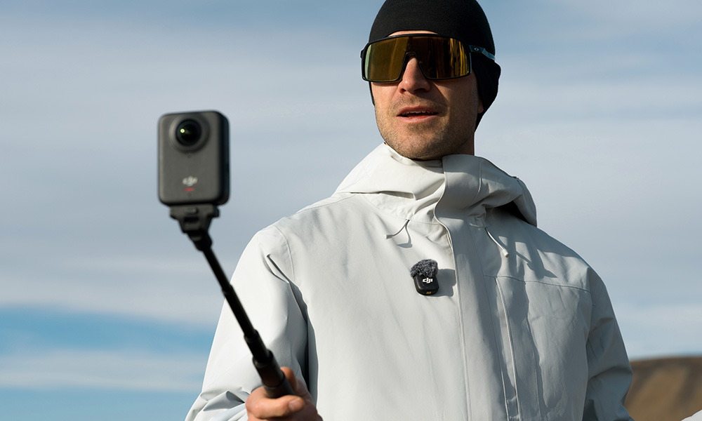 Mężczyzna w okularach przeciwsłonecznych i czapce trzymający kamerę na selfie sticku. Ma na sobie szarą kurtkę. Błękitne niebo w tle.