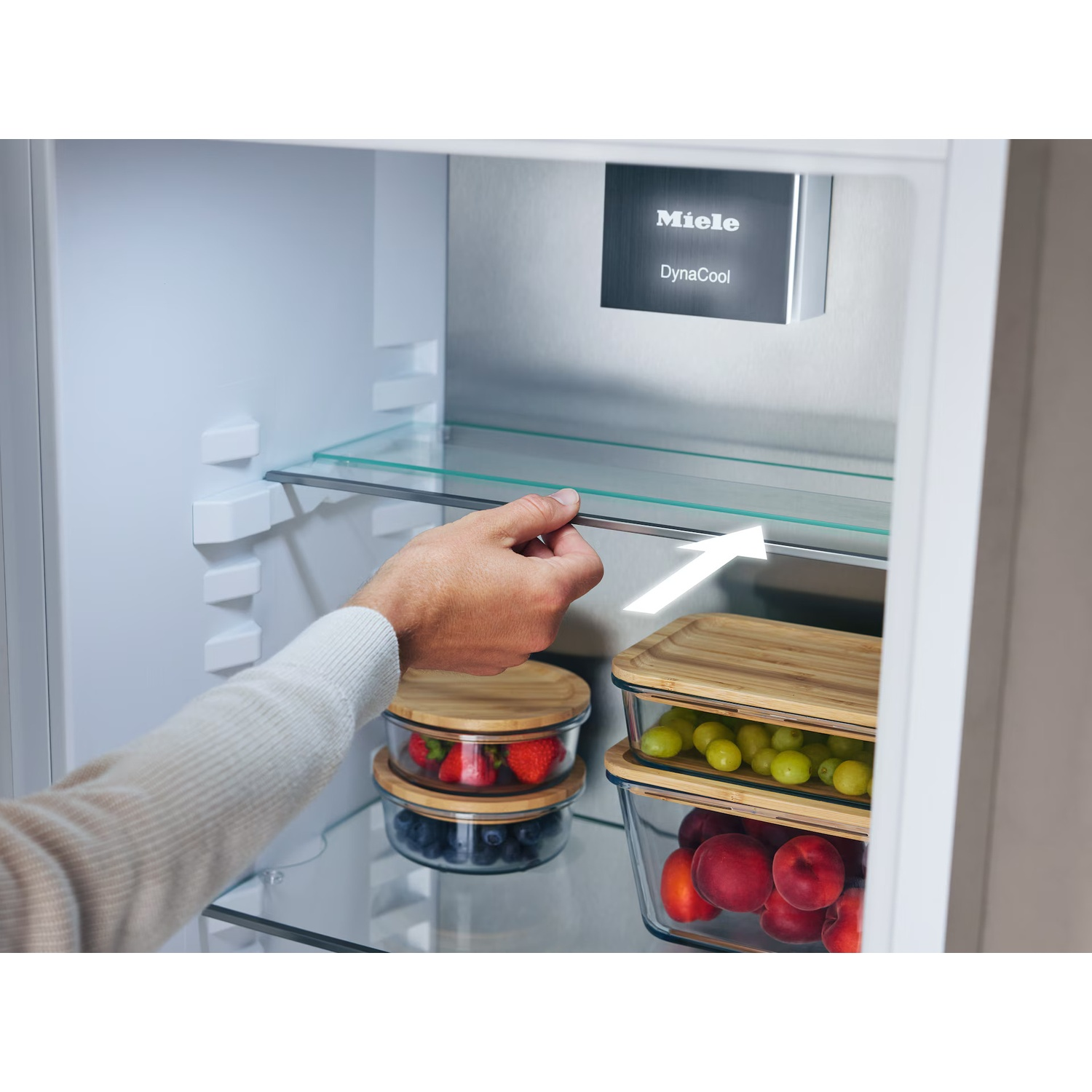 Een hand trekt een glazen plank in een Miele koelkast. Containers met fruit zoals aardbeien, bosbessen, druiven en perziken zijn zichtbaar.
