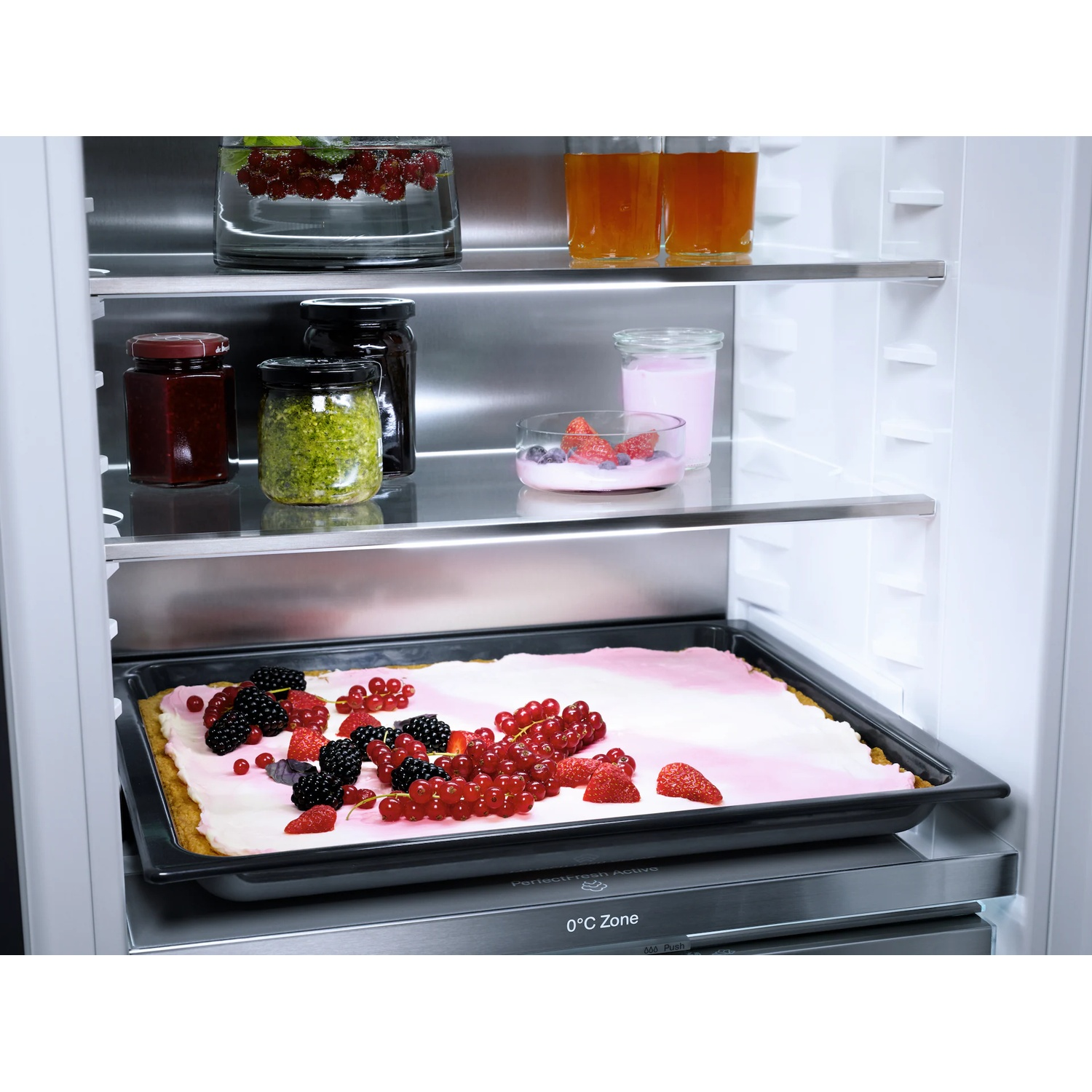 Een koelkast met diverse items: dessert met fruit, bessen, potten jam en pesto, yoghurt en sap.