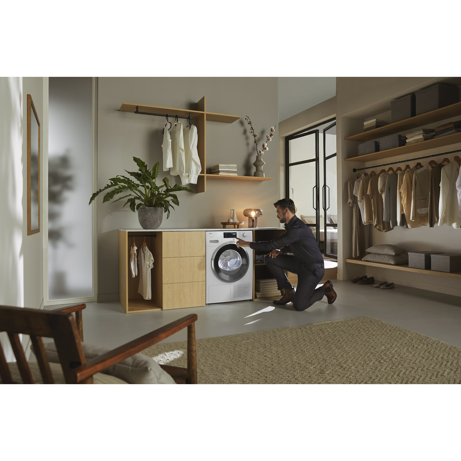 A man kneels by a washing machine in a modern laundry room. Clothes hang on shelves. A plant and a wooden chair are visible.