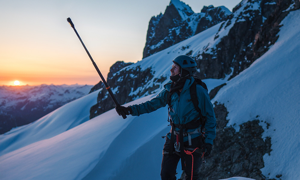 Osoba w zimowym stroju stoi na ośnieżonej górze, trzymając kij. W tle widoczny jest zachód słońca, z ośnieżonymi górami.