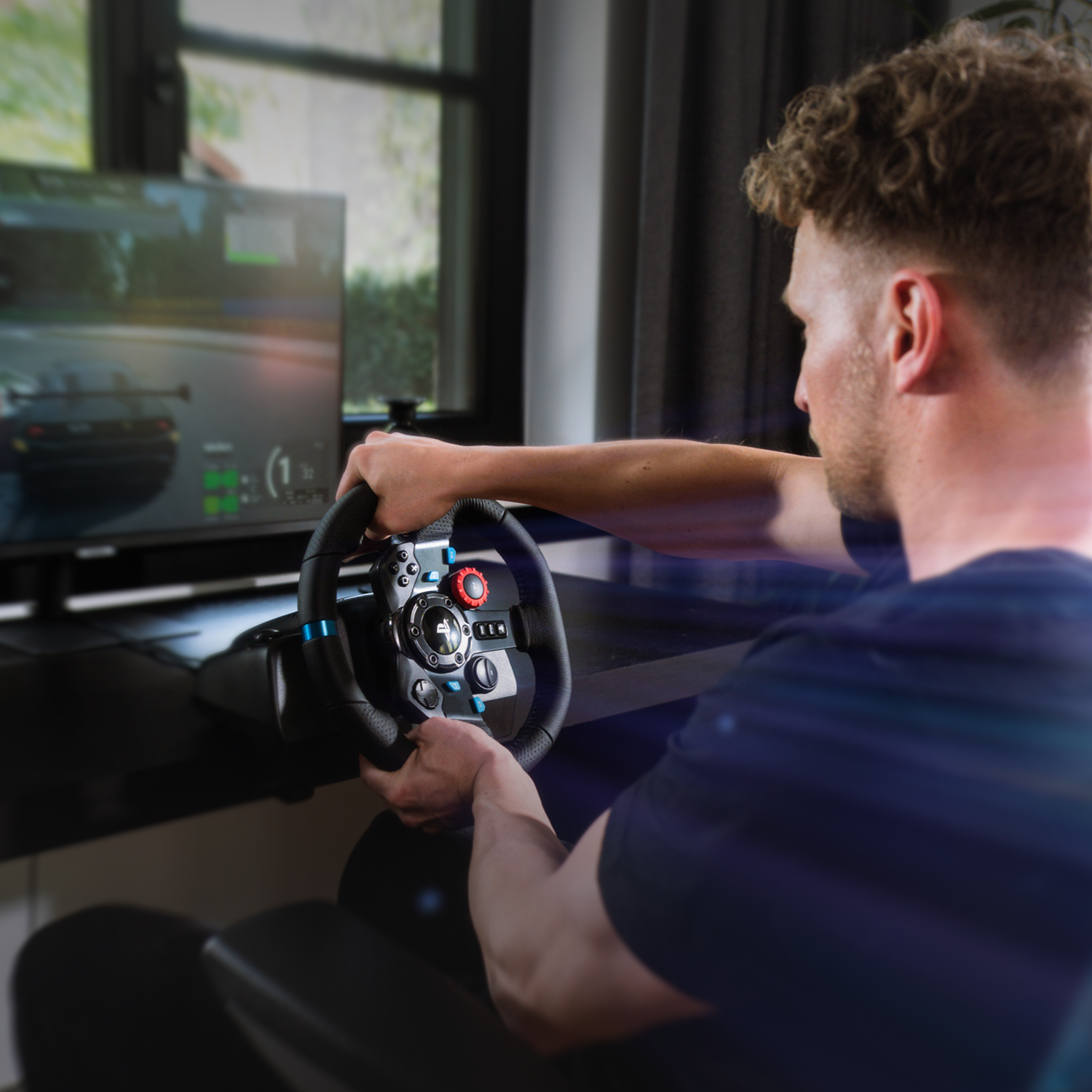 Un hombre juega un juego de carreras usando un volante. Una pantalla muestra la vista del juego. El hombre lleva una camisa azul.