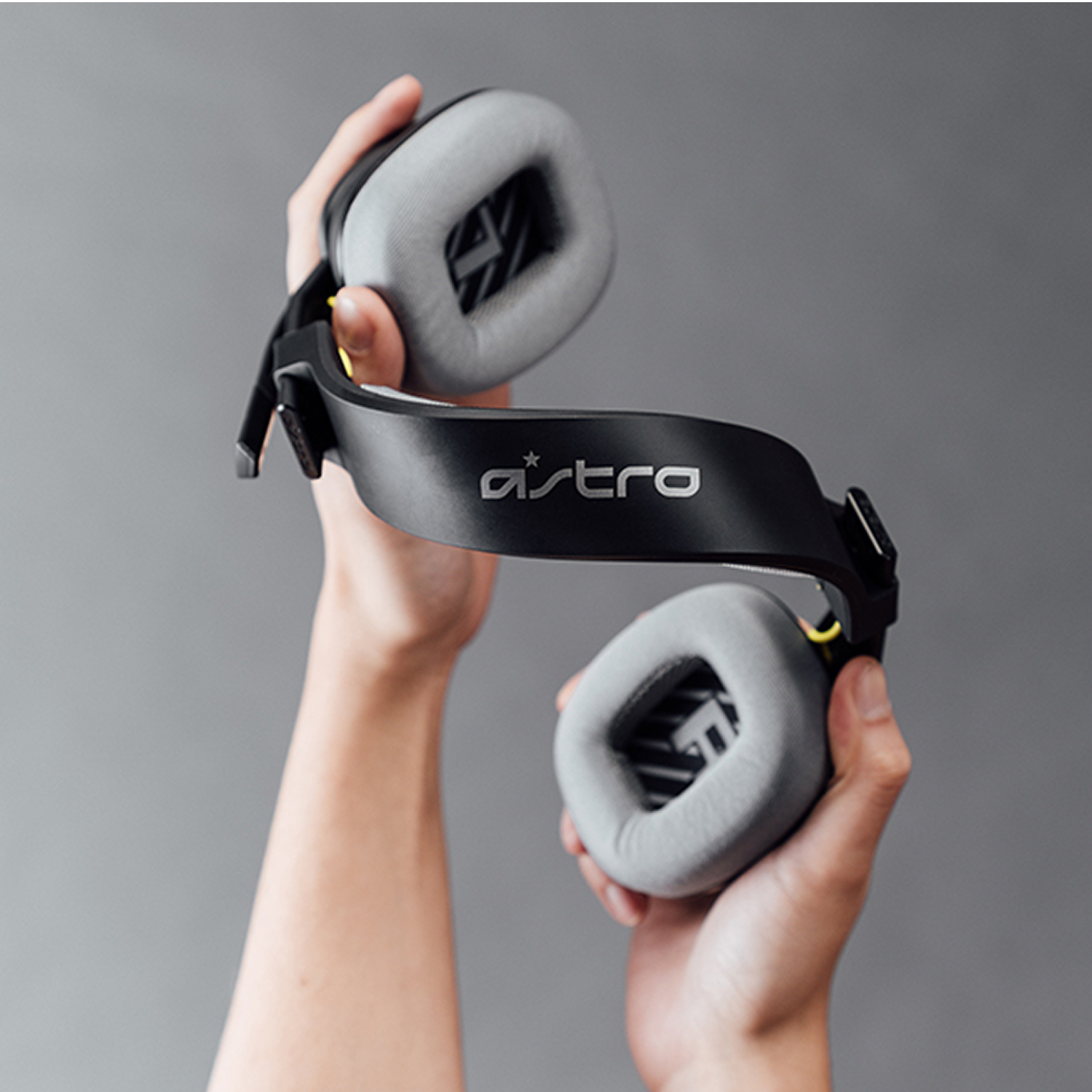 Manos sosteniendo auriculares Astro negros con almohadillas grises sobre un fondo gris. Los auriculares tienen el logo de la marca.