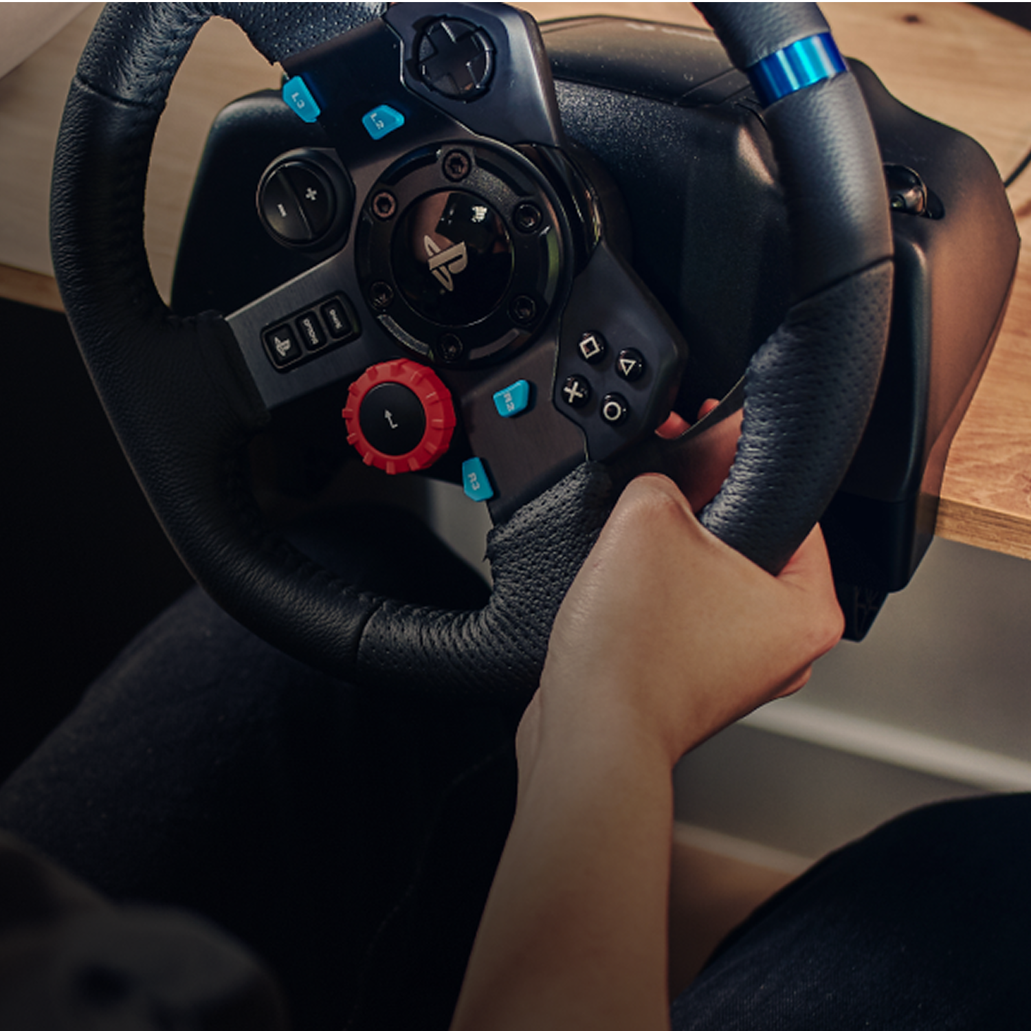 Una persona sostiene un volante negro, con botones azules y rojos. El fondo muestra una superficie de madera y la ropa de la persona.