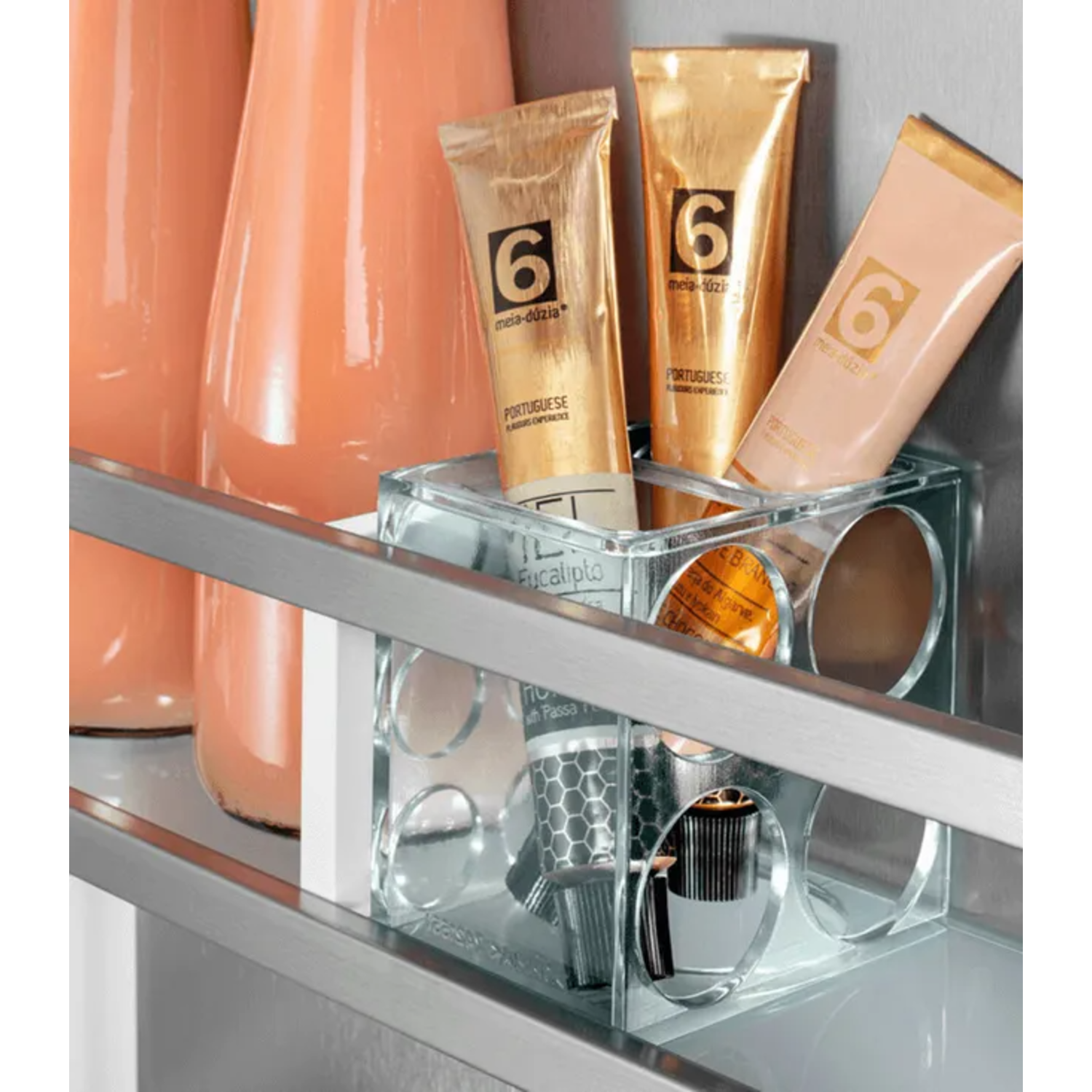 Three tubes of cosmetics in a clear container, next to a light orange liquid in bottles. The tubes have a gold color.