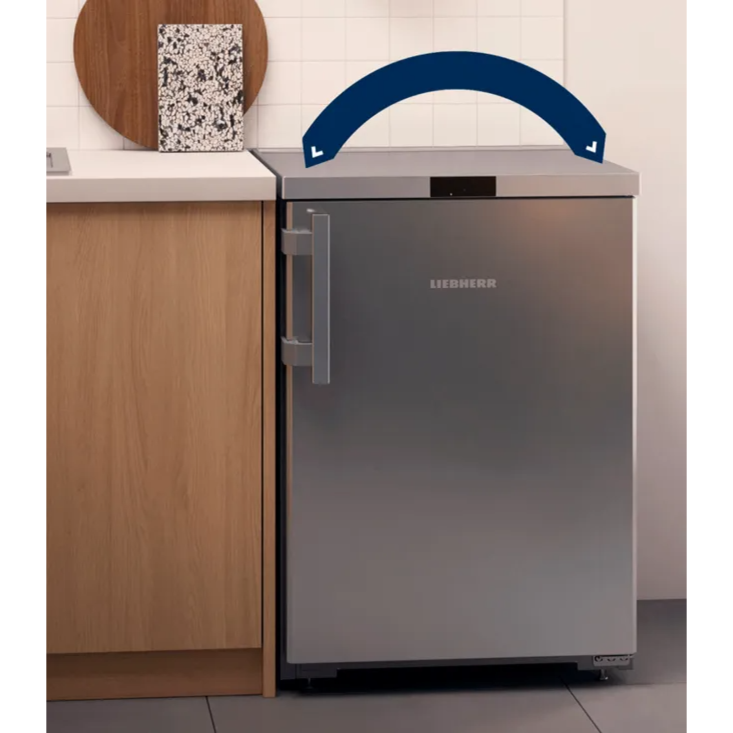 A silver refrigerator stands beside a wooden cabinet. A cutting board and a decorative item rest on the counter above.