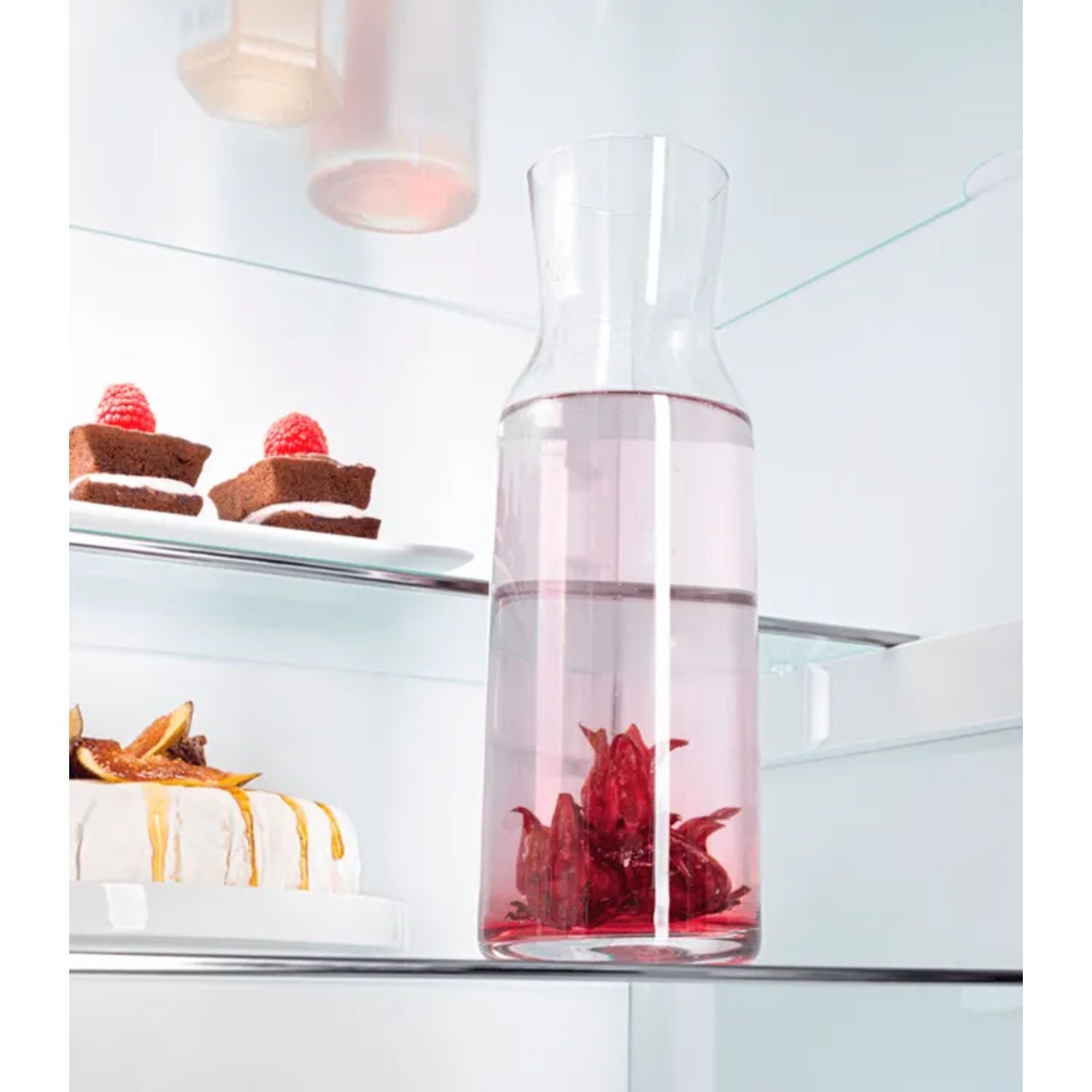 A refrigerator shelf displays a pitcher of pink liquid with red flowers, two small cakes with raspberries, and a cake.