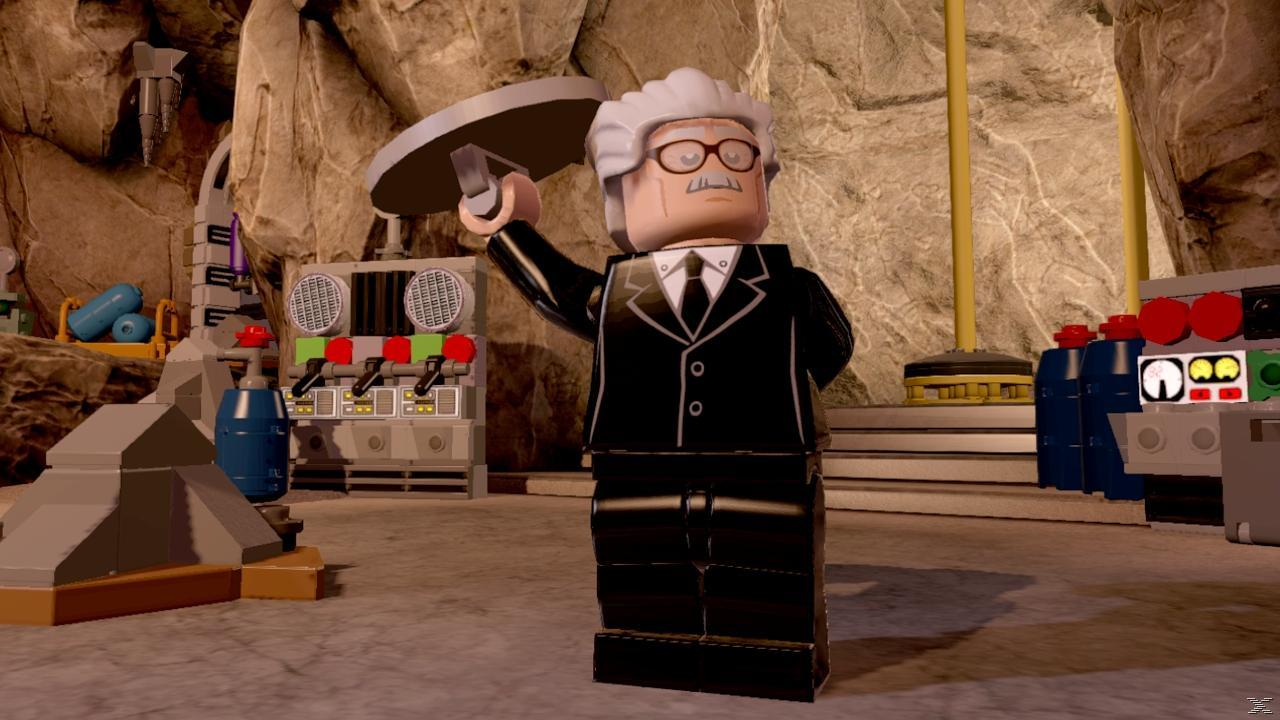 Lego Alfred w czarnym garniturze, trzymający srebrny przedmiot. Scena w jaskini z maszynami i kamiennymi ścianami.