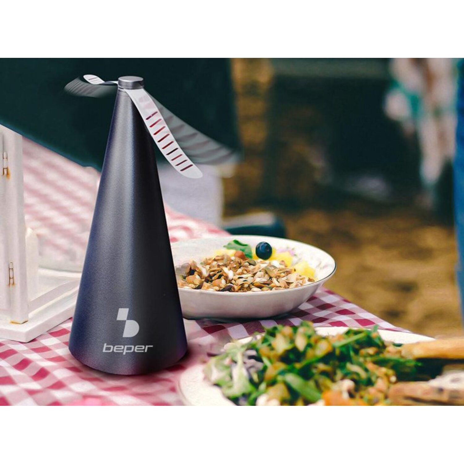 Een zwarte, kegelvormige Beper-ventilator staat op een rood-geruite tafel. Salade en een kom met eten staan ernaast. De achtergrond is wazig.