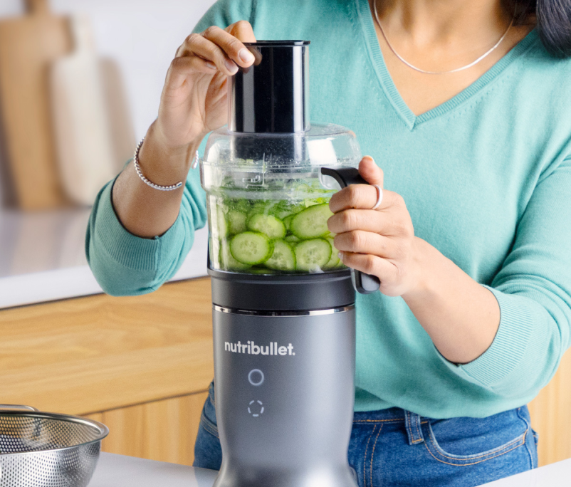Eine Person verwendet einen Nutribullet, um Gurken zu schneiden. Die Maschine ist grau, und die Person trägt einen türkisblauen Pullover und Jeans.