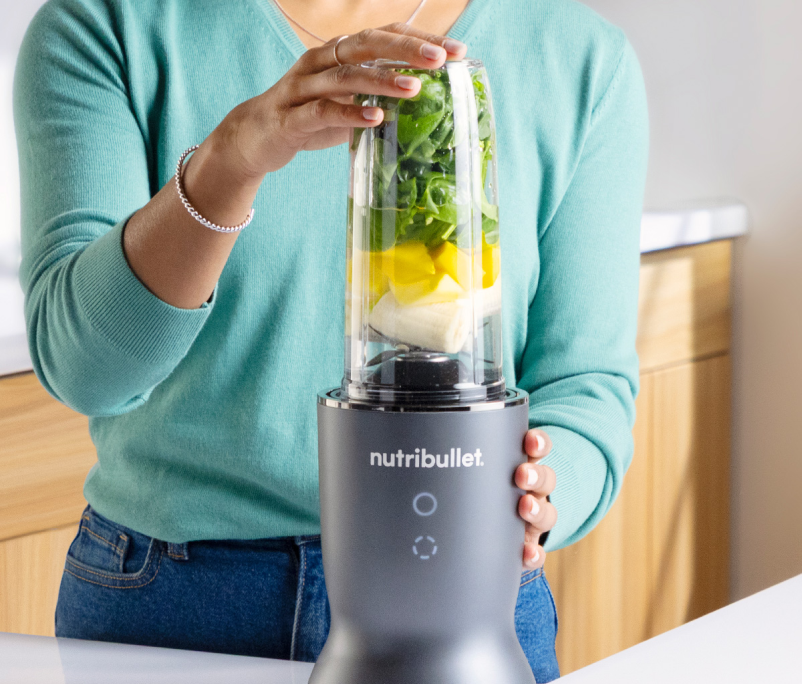 Eine Person hält einen Nutribullet-Mixer mit Zutaten darin. Der Mixer ist grau. Die Person trägt einen türkisfarbenen Pullover und Jeans.
