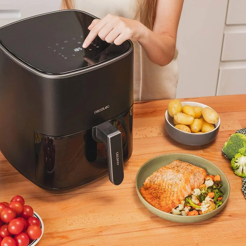 Una persona usa una freidora de aire negra. En la mesa hay cuencos con tomates, patatas, salmón con verduras y brócoli.