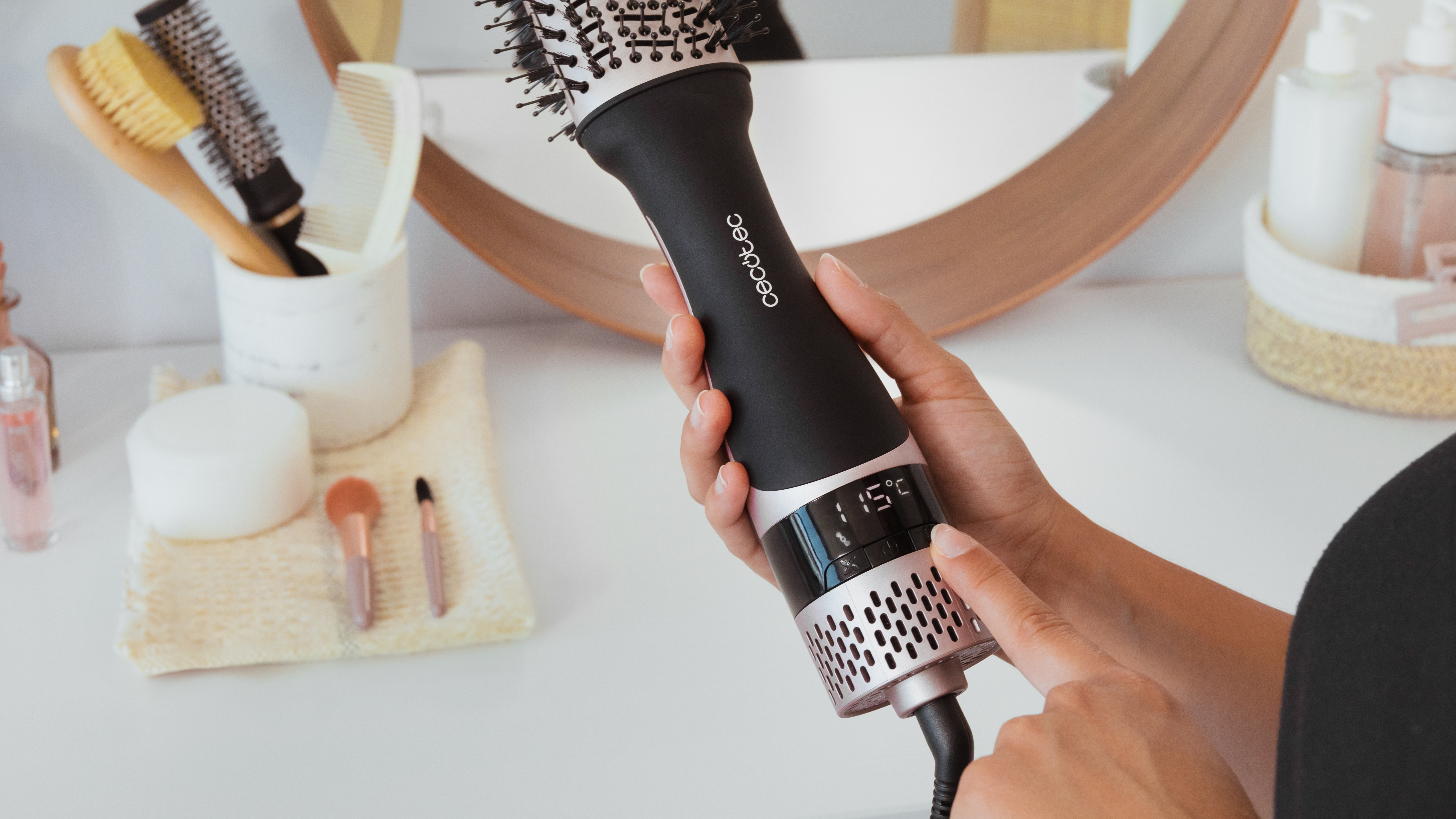 Una persona sostiene un secador de pelo negro y rosa, con una pantalla digital que muestra 115 grados. Otros accesorios para el cabello están sobre la mesa.