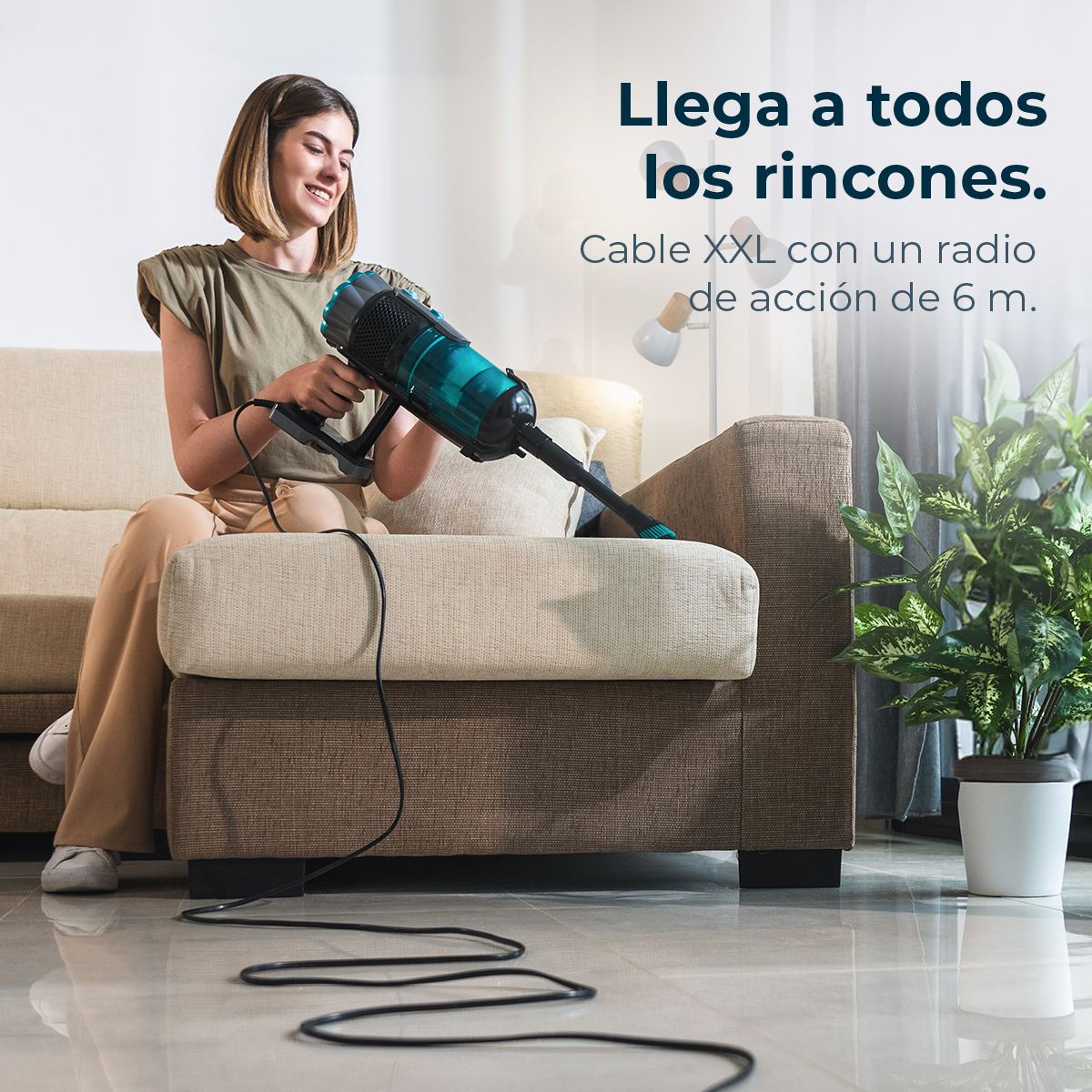 Una mujer aspira un sofá con una aspiradora verde y negra. Una planta en maceta está a la derecha. El texto dice: 'Llega a todos los rincones'.