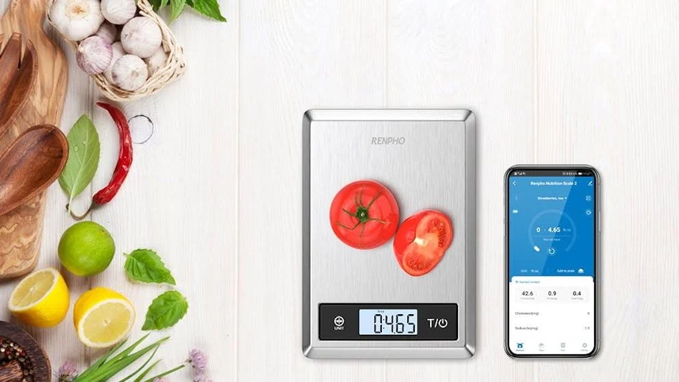Una báscula de cocina digital con tomates, un smartphone con datos y varias frutas y verduras sobre una superficie de madera.