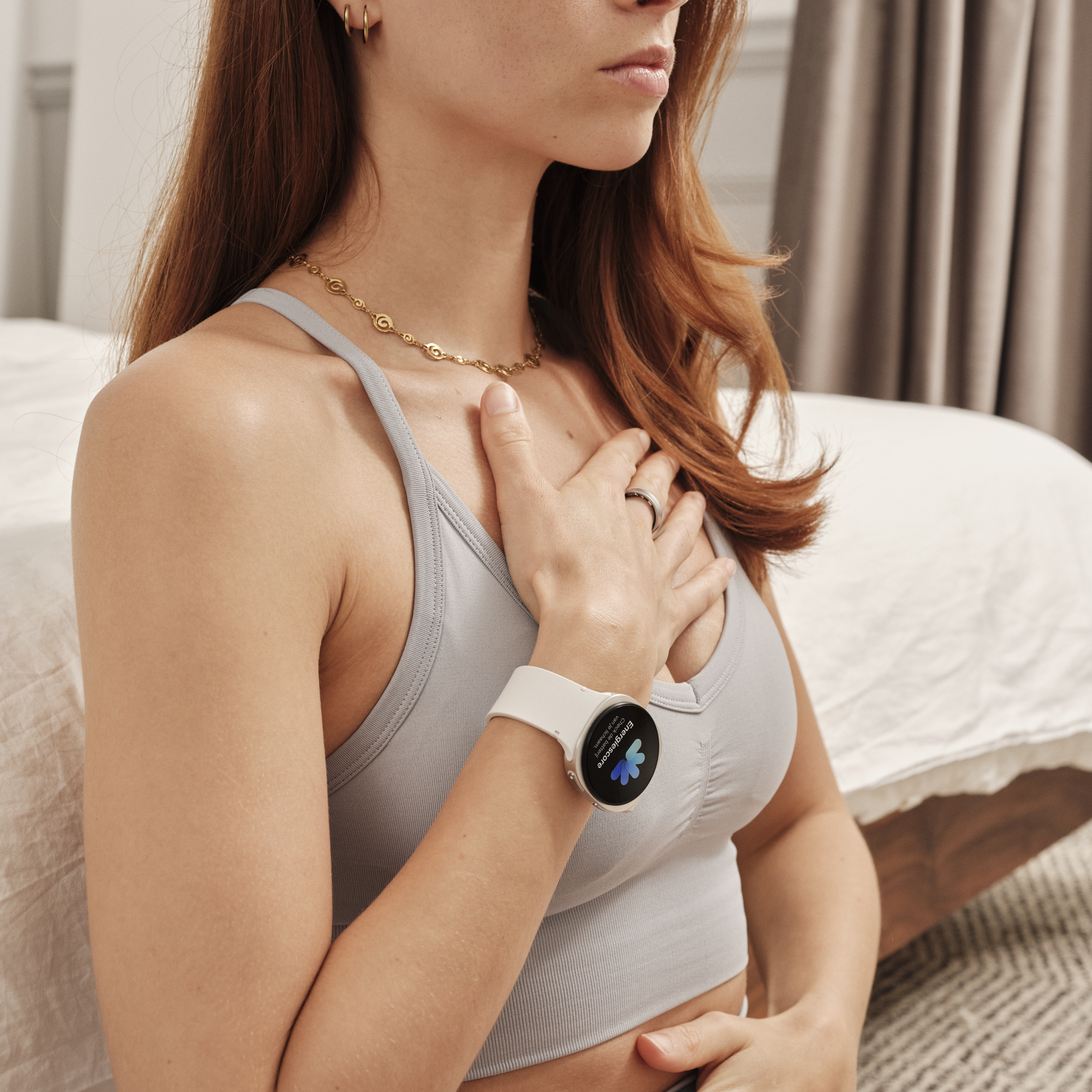 Woman wearing a smartwatch, touching chest. She has red hair, gold jewelry. Background includes a bed, curtains, and a rug.
