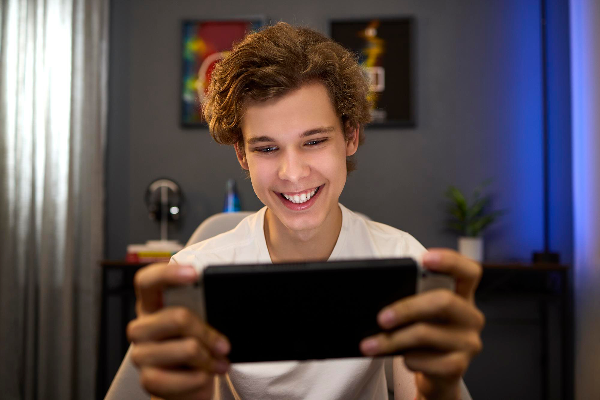 Ein junger Mann lächelt, während er ein schwarzes Smartphone hält. Er sitzt auf einem Stuhl. Der Hintergrund umfasst Kunst, Kopfhörer und eine Pflanze.
