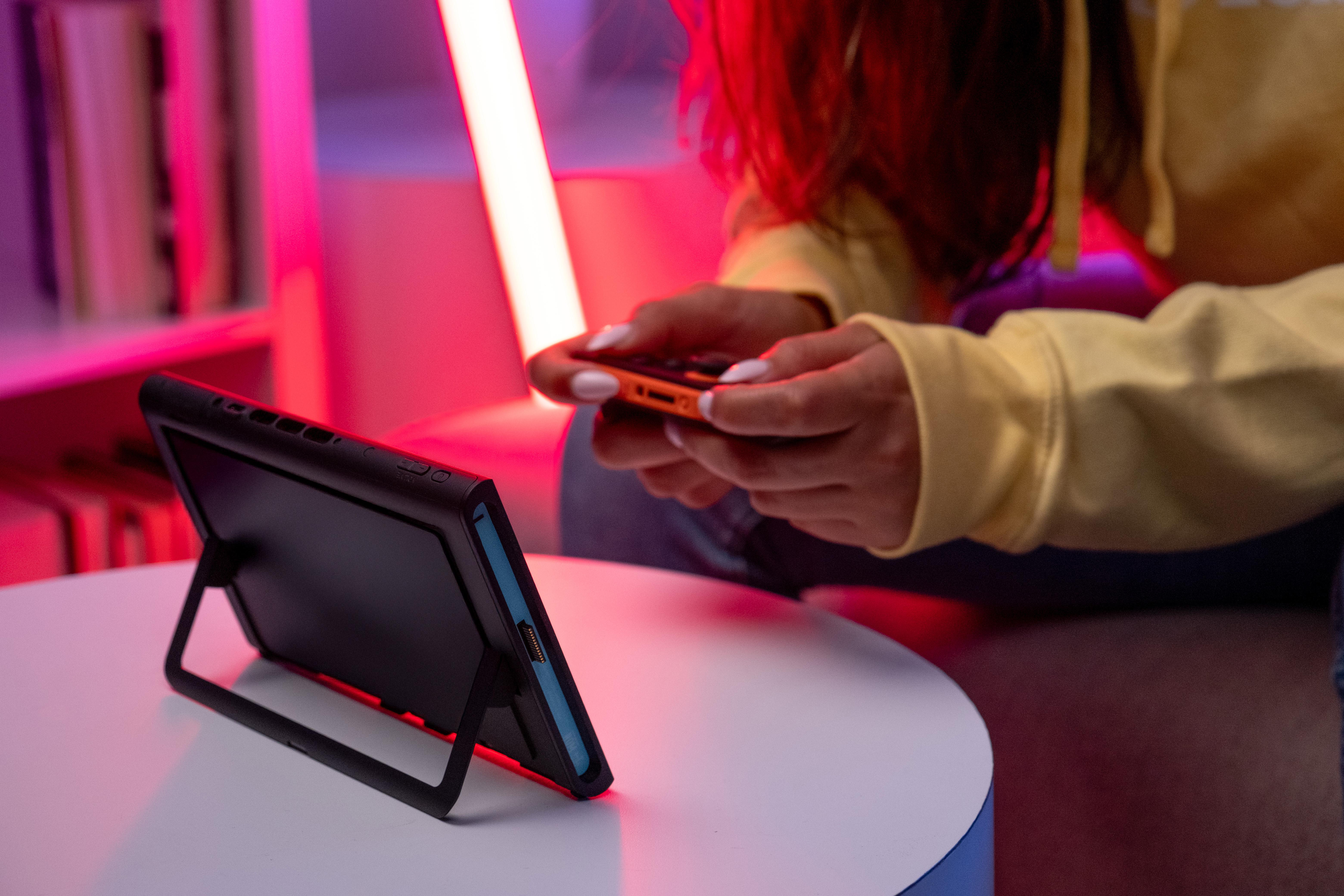 Eine Person hält eine Spielkonsole auf einem Tisch. Der Hintergrund wird von rosa und roten Lichtern beleuchtet.