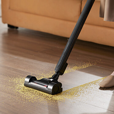 Czarny odkurzacz czyści żółte cząsteczki na drewnianej podłodze, w pobliżu beżowej sofy.