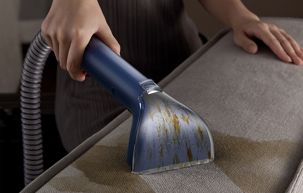 Osoba używa niebieskiego i przezroczystego narzędzia czyszczącego na szarej sofie, usuwając plamy po płynie. Ręka trzyma narzędzie.