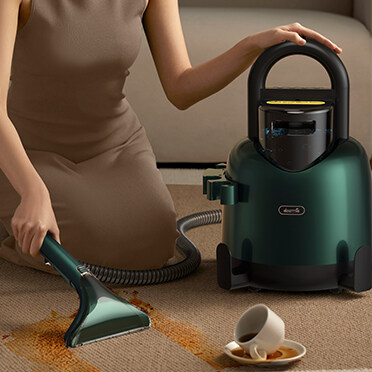 Kobieta używa zielonego odkurzacza na dywanie z rozlaną filiżanką kawy w pobliżu.