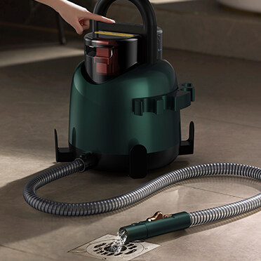 Ręka dotyka ciemnozielonego odkurzacza. Wąż rozciąga się do odpływu, z którego wypływa woda.